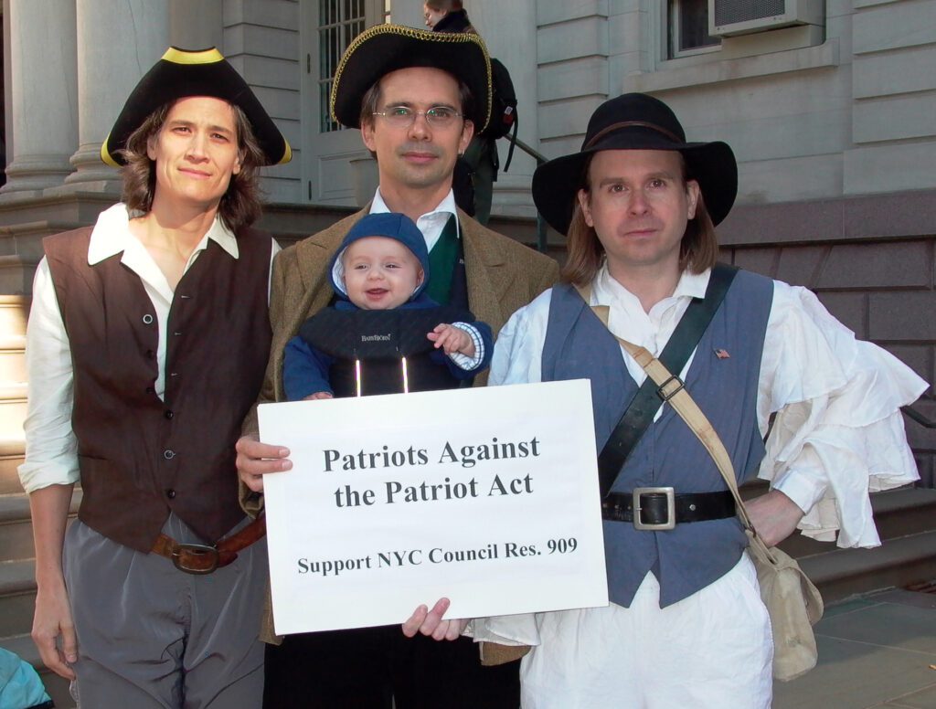 Demonstrators dressed as 18th Century American Revolutionaries demonstrate in front of City Hall in 2003. They favored a resolution before the New York City Council supporting restoration of civil liberties curtailed by the government since Sept. 11, 2001. Despite the City Council passing the resolution in 2004--and more than 250 other cities and three states, provisions of the Patriot Act remain in effect in 2022. Objections are still relevant. See https://www.aclu.org/other/surveillance-under-usapatriot-act. Left to right: Kelly Moore, Stephen Duncombe, Sydney Railla-Duncombe (aged 4 months), Bob Lesko.  Photo by Pat Arnow